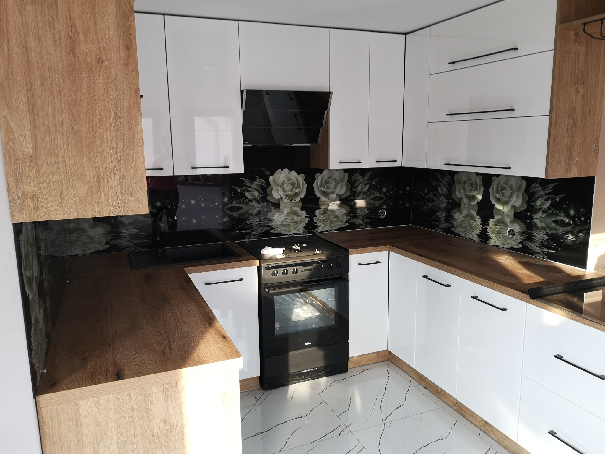 Nowoczesna kuchnia z białymi, lakierowanymi szafkami, drewnianymi blatami i czarnym panelem szklanym z motywem białych róż nad blatem roboczym i płytą grzewczą.