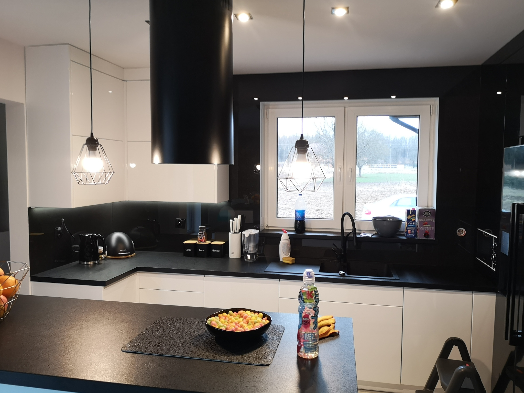 Nowoczesna kuchnia z białymi szafkami i czarnymi blatami, z widocznym oknem i elementami dekoracyjnymi, takimi jak lampy wiszące o geometrycznym kształcie i kolorowe słodycze w misce na wyspie.