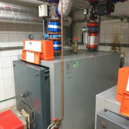 Tomasz Jagodziński AQUATERM - Kocioł grzewczy Viessmann Vitoplex 100 w kotłowni z widocznymi rurami, zaworami i elementami instalacji. Rury izolowane folią aluminiową biegną wzdłuż sufitu, a na ścianie widać białe płytki.