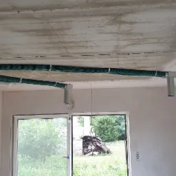 Zielone rury wentylacyjne przymocowane do sufitu w surowym pomieszczeniu, widoczne okna i drzwi balkonowe, na ścianie wisi młotek.