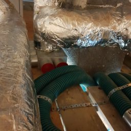 Tomasz Jagodziński AQUATERM - Fragment poddasza z widocznymi izolowanymi przewodami wentylacyjnymi i elementami systemu rekuperacji, w tym zielonymi rurami karbowanymi i srebrną folią izolacyjną na kanałach.