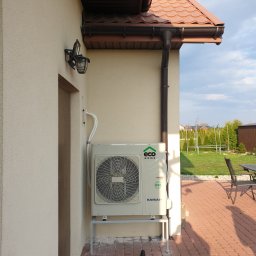 Tomasz Jagodziński AQUATERM - Zewnętrzna jednostka pompy ciepła marki Kaisai zamontowana na metalowym stelażu przy ścianie budynku z brązowym dachem i rynną, widoczna instalacja rur, obok drzwi wejściowe i lampa zewnętrzna,...