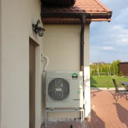 Zewnętrzna jednostka pompy ciepła marki Kaisai zamontowana na metalowym stelażu przy ścianie budynku z brązowym dachem i rynną, widoczna instalacja rur, obok drzwi wejściowe i lampa zewnętrzna,...