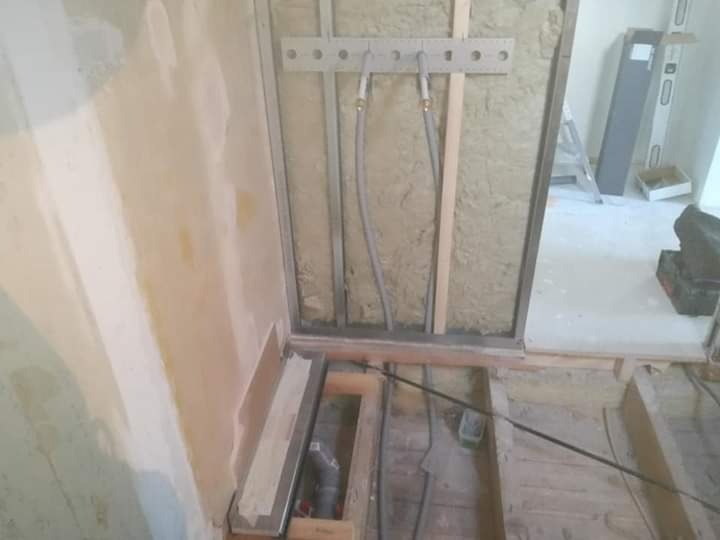 Wnętrze łazienki w trakcie remontu, widoczne odsłonięte rury instalacji wodnej zamocowane do ściany z izolacją, surowa podłoga z drewnianych belek oraz częściowo otynkowana ściana w tle.