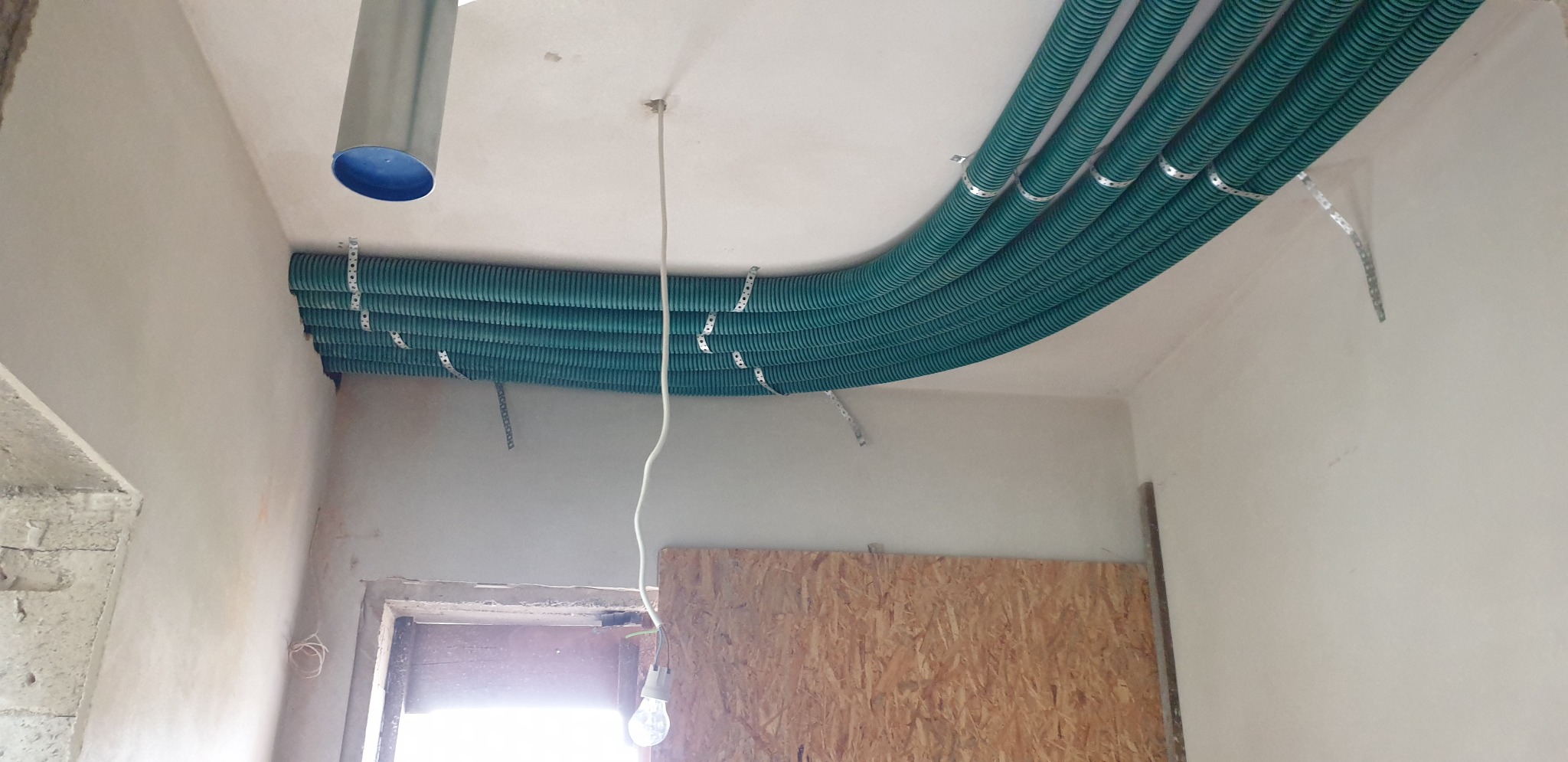 Równolegle ułożone, zielone, karbowane rury wentylacyjne pod sufitem, mocowane metalowymi obejmami, widoczny fragment surowego wnętrza w trakcie prac wykończeniowych.