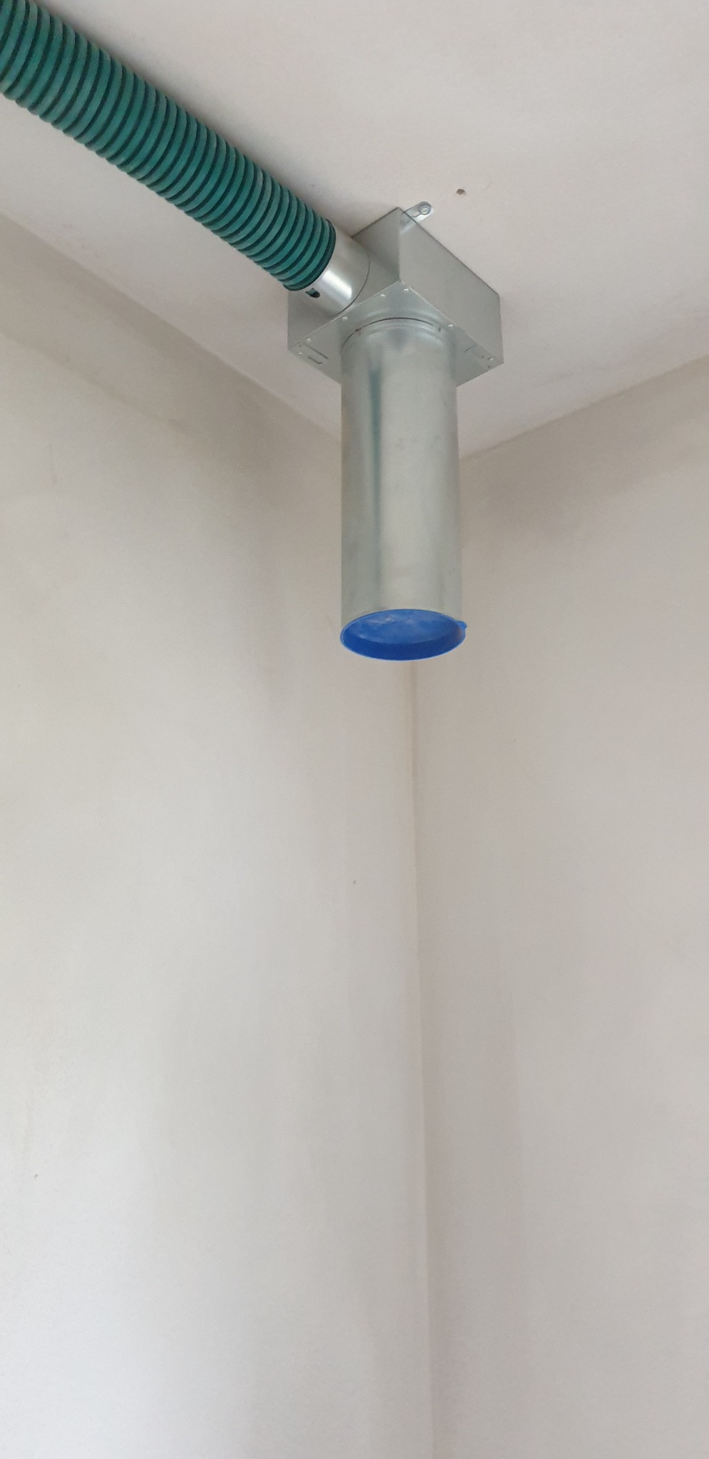 Szary, metalowy kanał wentylacyjny z niebieską zaślepką, przymocowany do sufitu w rogu pomieszczenia, połączony z zielonym, elastycznym przewodem wentylacyjnym.