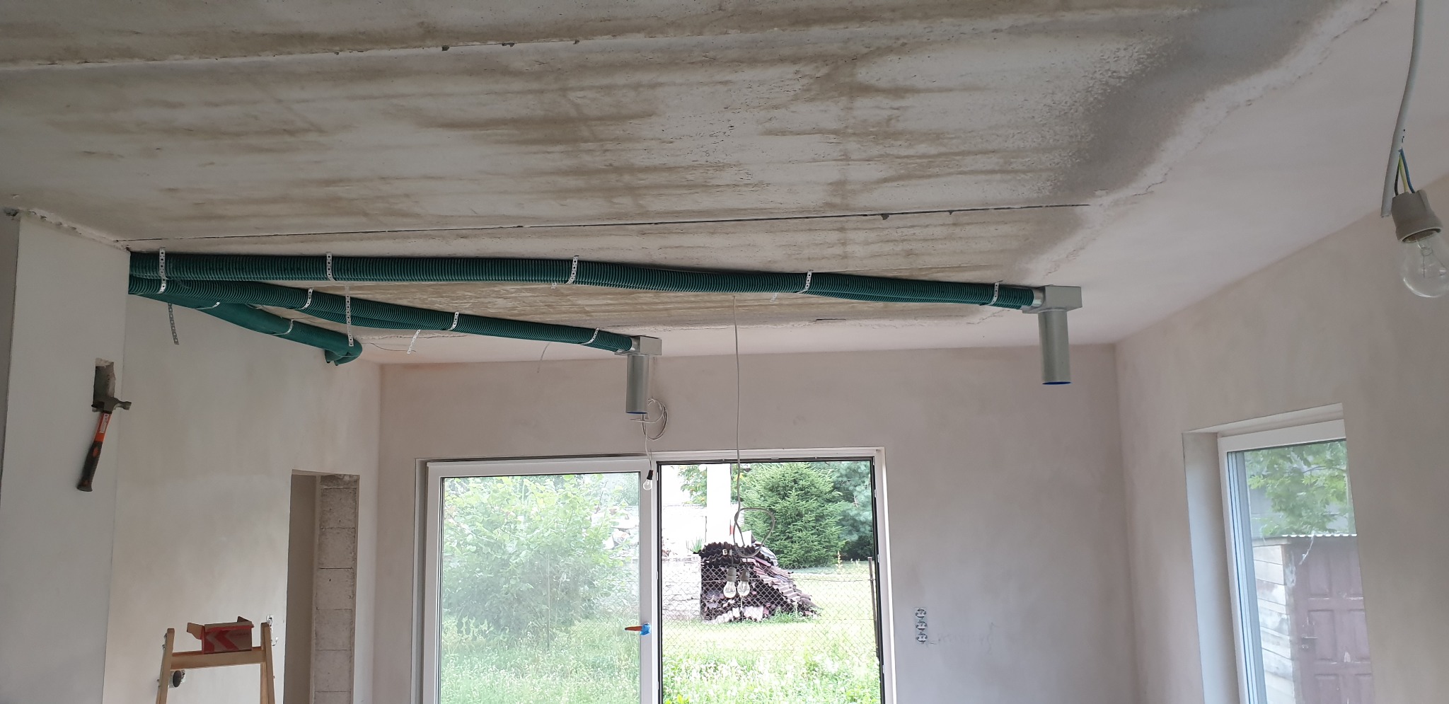 Zielone rury wentylacyjne przymocowane do sufitu w surowym pomieszczeniu, widoczne okna i drzwi balkonowe, na ścianie wisi młotek.