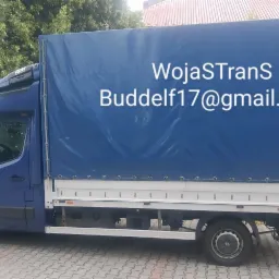Niebieski bus z plandeką zaparkowany na chodniku, z widocznym logo firmy 'WojaSTrans' i adresem email 'Buddelf17@gmail.com' na plandece.