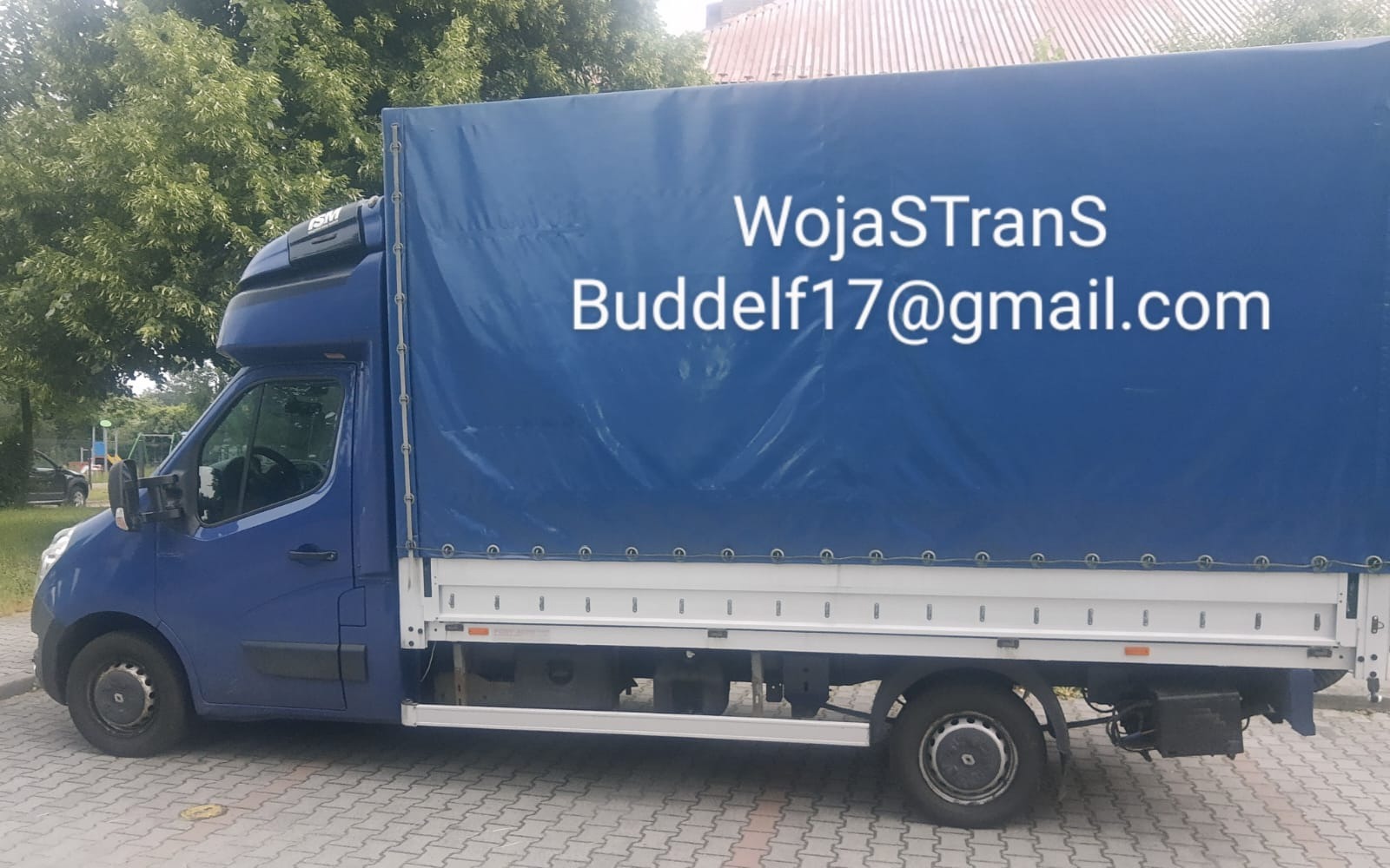 Niebieski bus z plandeką zaparkowany na chodniku, z widocznym logo firmy 'WojaSTrans' i adresem email 'Buddelf17@gmail.com' na plandece.