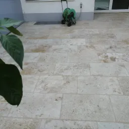 Jasny taras wyłożony prostokątnymi płytami z naturalnego kamienia trawertynu, z widocznym fragmentem rośliny w lewym dolnym rogu oraz zwiniętym wężem ogrodowym na wózku w tle.