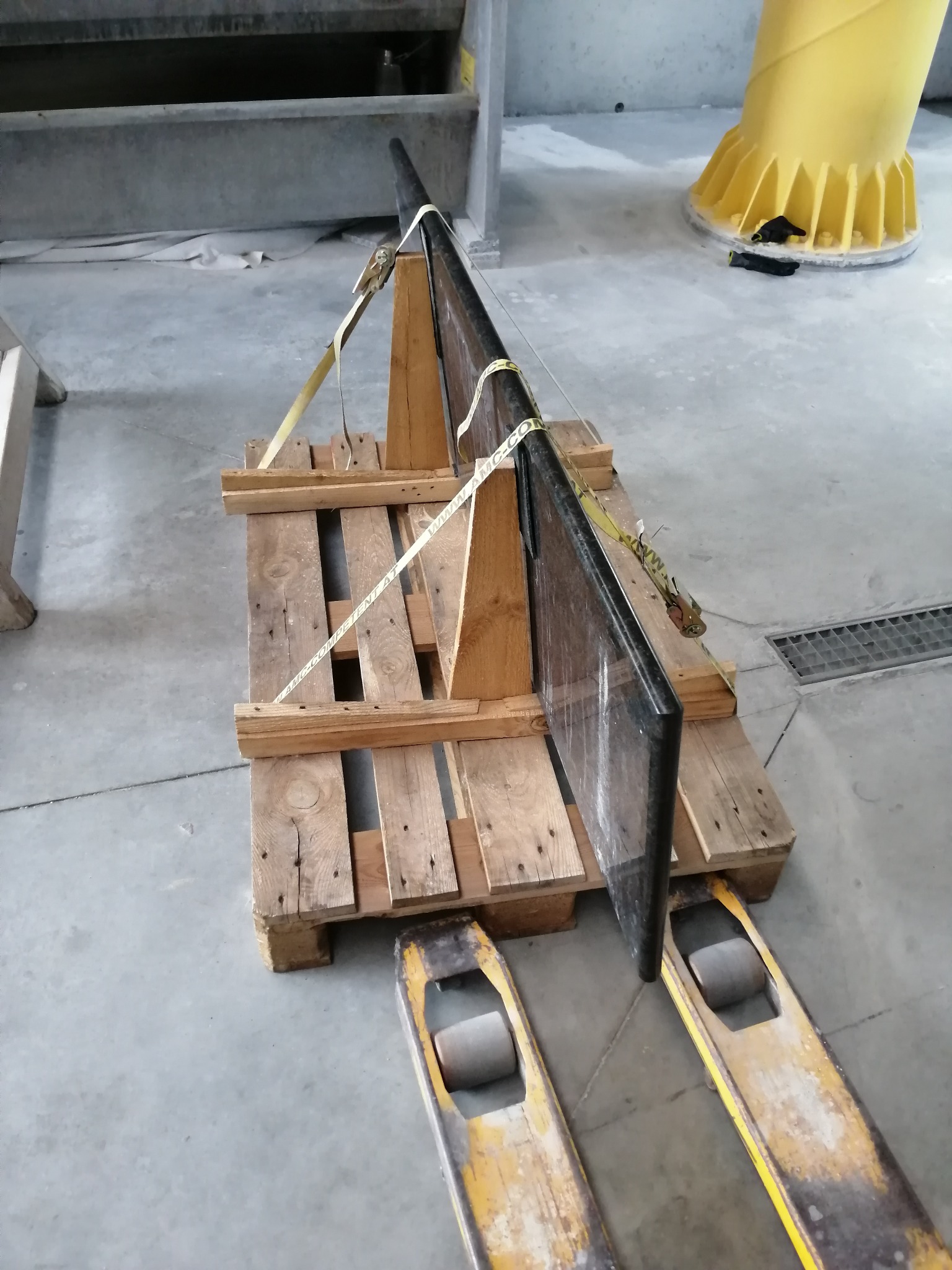 Czarny, polerowany kamienny element, zabezpieczony pasami transportowymi na drewnianej palecie, gotowy do transportu w hali produkcyjnej.