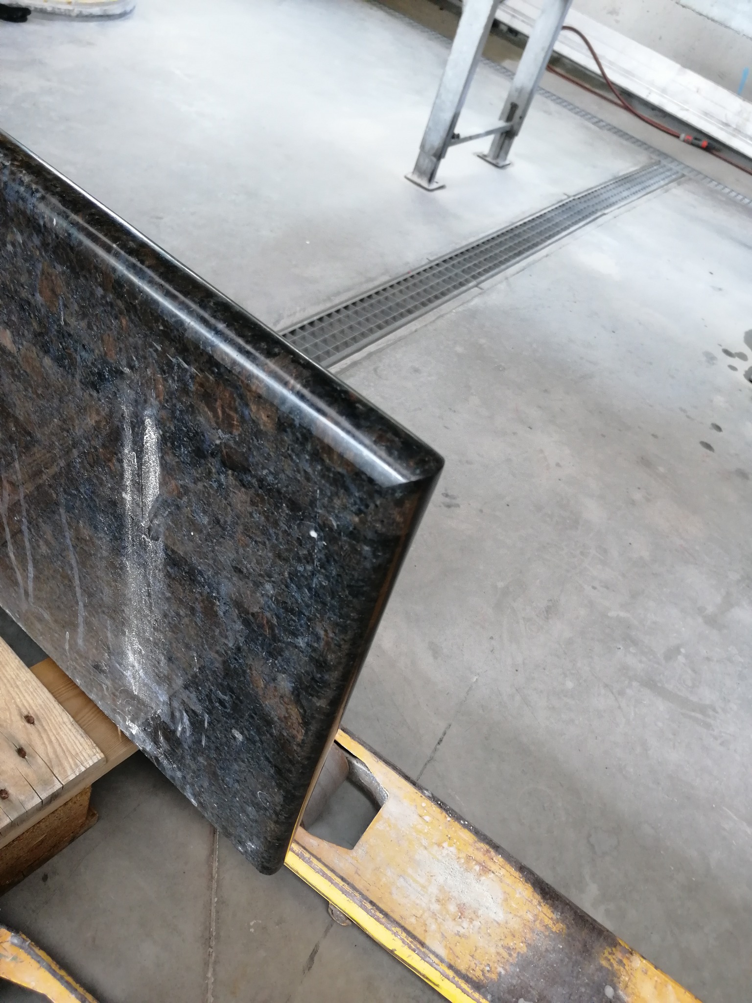 Ciemny, polerowany blat kamienny z zaokrąglonym brzegiem, oparty na drewnianej palecie i żółtym wózku transportowym w warsztacie kamieniarskim.