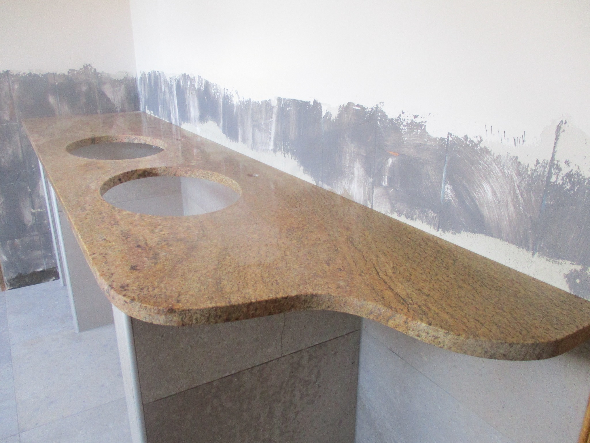 Blat łazienkowy z brązowego granitu z dwoma okrągłymi otworami na umywalki, zamontowany na szafkach w trakcie remontu, ściana przygotowana pod malowanie.