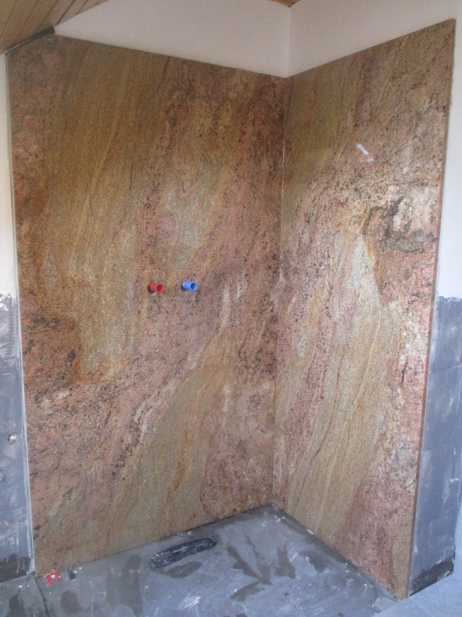 Wyłożona brązowo-różowym kamieniem ściana kabiny prysznicowej z widocznymi przyłączami ciepłej i zimnej wody oraz odpływem w podłodze, w trakcie prac wykończeniowych.