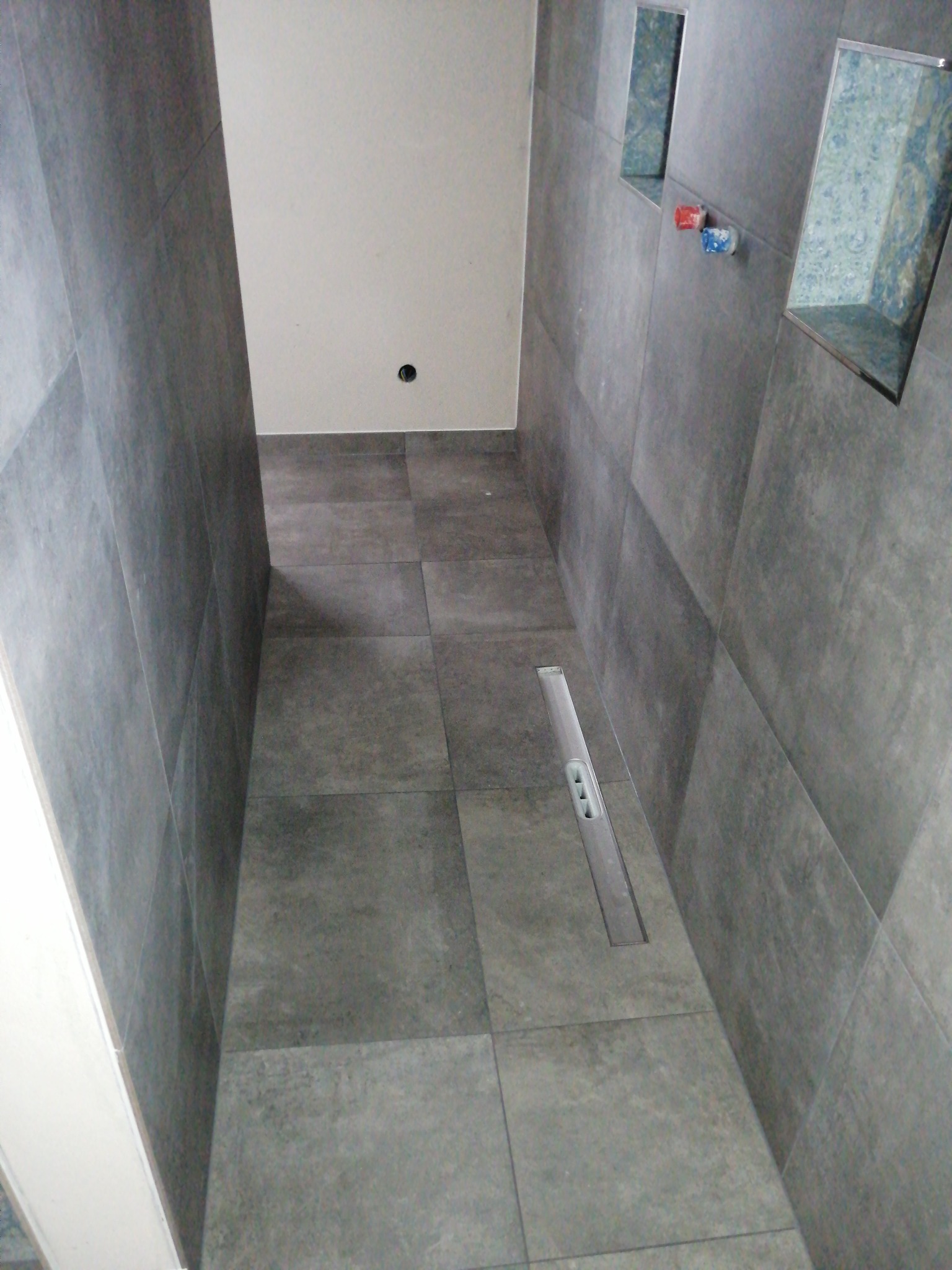 Wykończona łazienka z szarymi, matowymi płytkami, liniowym odpływem podłogowym i wnękami w ścianie z dekoracyjnym wykończeniem.