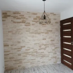 Ściana wykończona panelami imitującymi kamień, nowoczesna lampa wisząca i drewniane drzwi z białymi akcentami w jasnym pomieszczeniu.