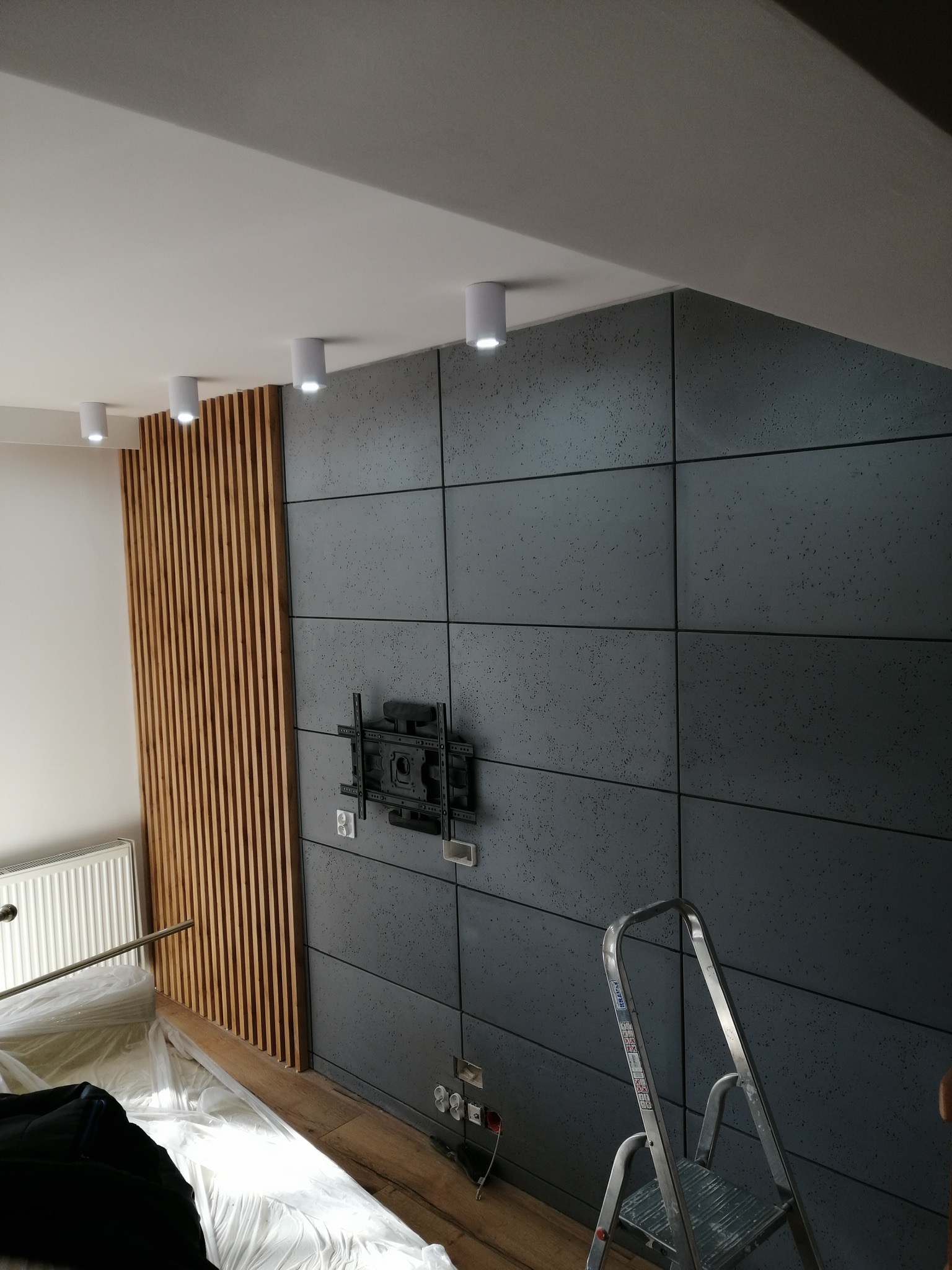 Wnętrze pokoju z dekoracyjną ścianą z szarych paneli i drewnianych listew, przygotowane do montażu telewizora, z widoczną drabiną i zabezpieczeniem podłogi folią.