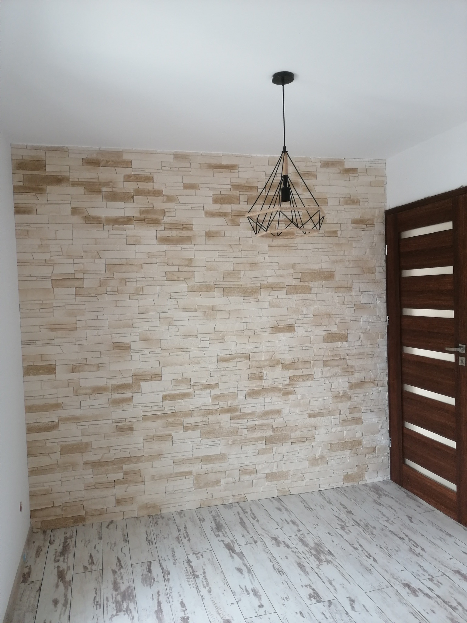 Ściana wykończona panelami imitującymi kamień, nowoczesna lampa wisząca i drewniane drzwi z białymi akcentami w jasnym pomieszczeniu.