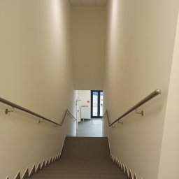Elita - Klatka schodowa z szarymi płytkami na schodach, białe ściany z metalowymi poręczami, widok z góry na dół, drzwi wejściowe na końcu korytarza.