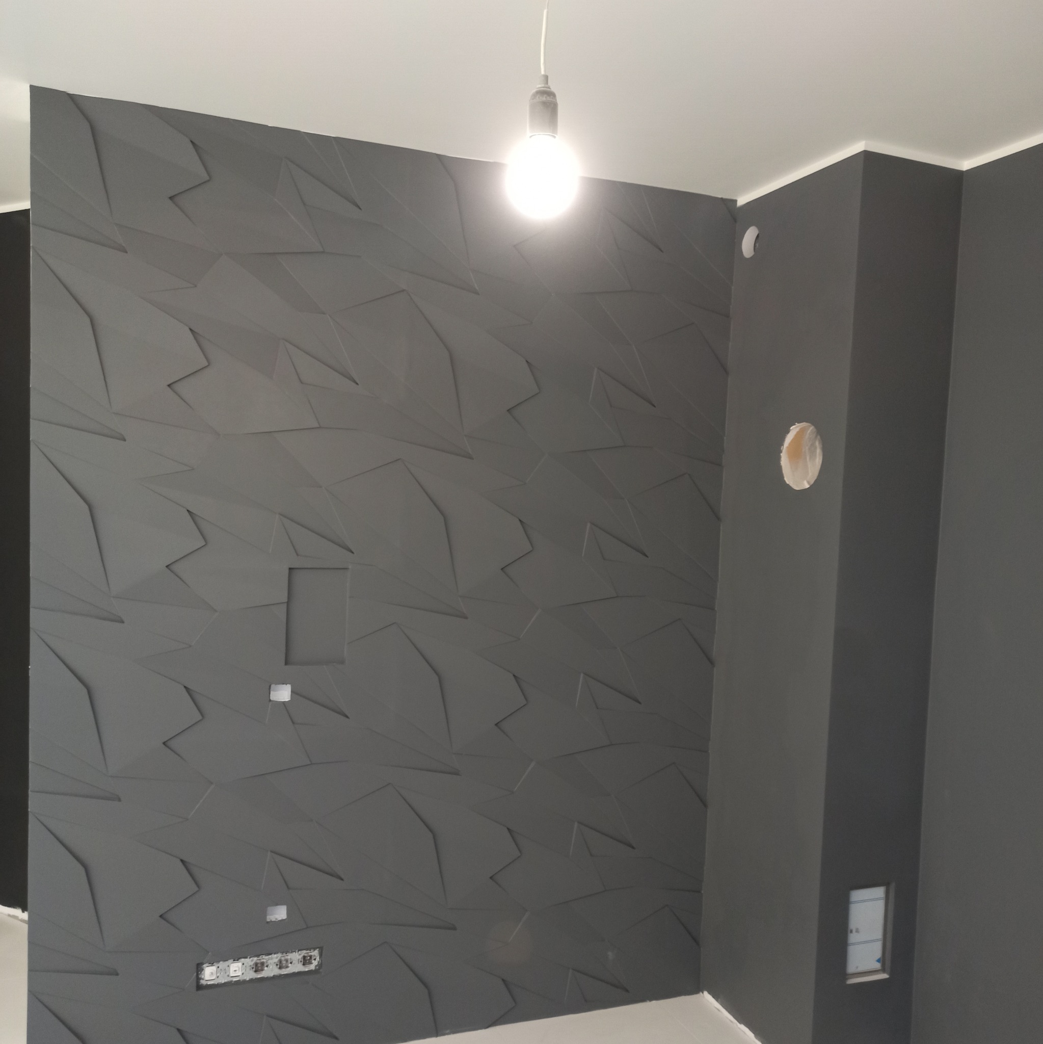 Szara ściana z geometrycznym wzorem 3D, widoczne otwory na gniazdka elektryczne i oświetlenie sufitowe