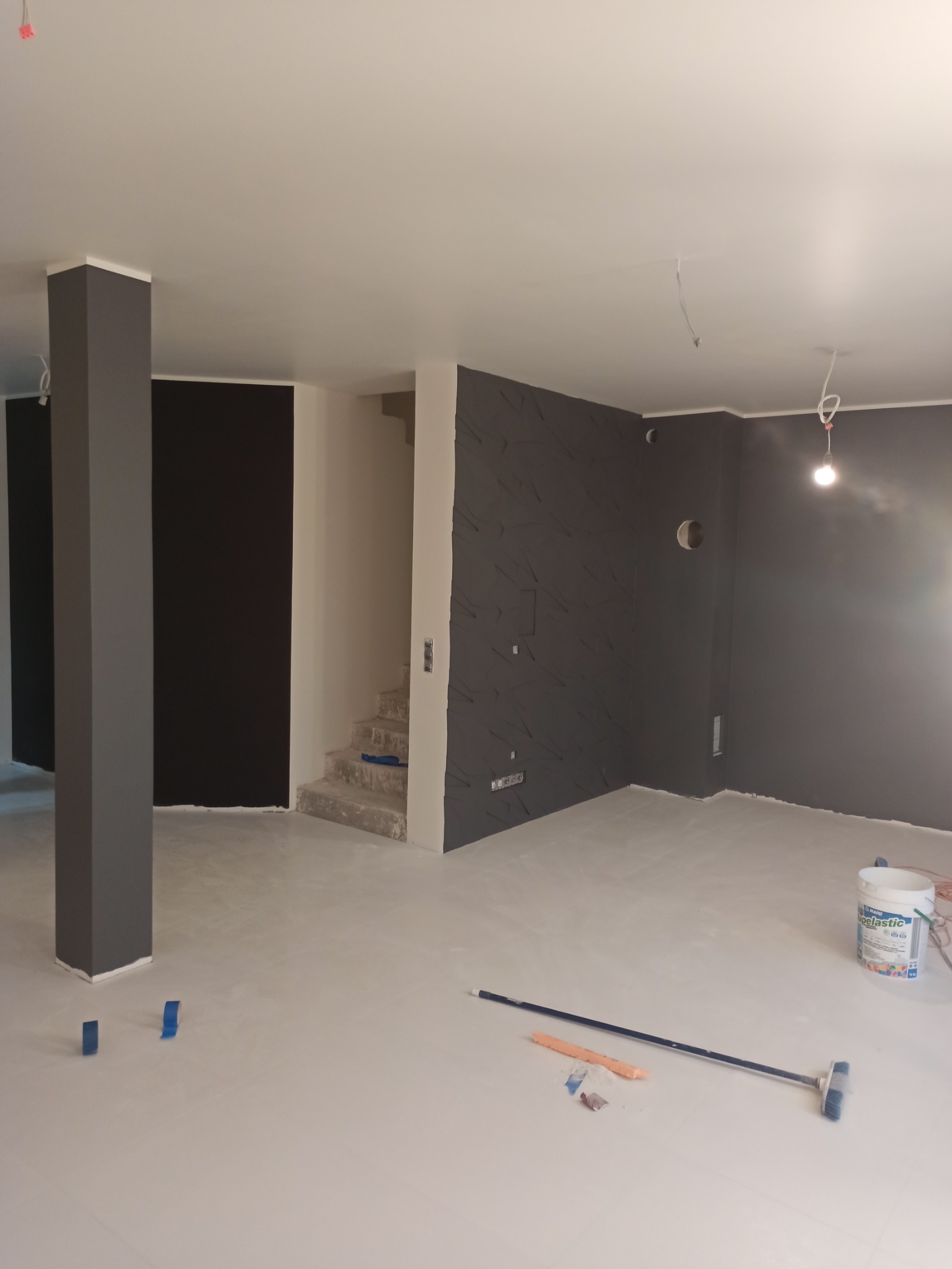 Wnętrze w trakcie prac wykończeniowych: szare ściany z dekoracyjną fakturą, przygotowane schody, zabezpieczona podłoga, widoczna puszka z masą szpachlową i narzędzia malarskie.