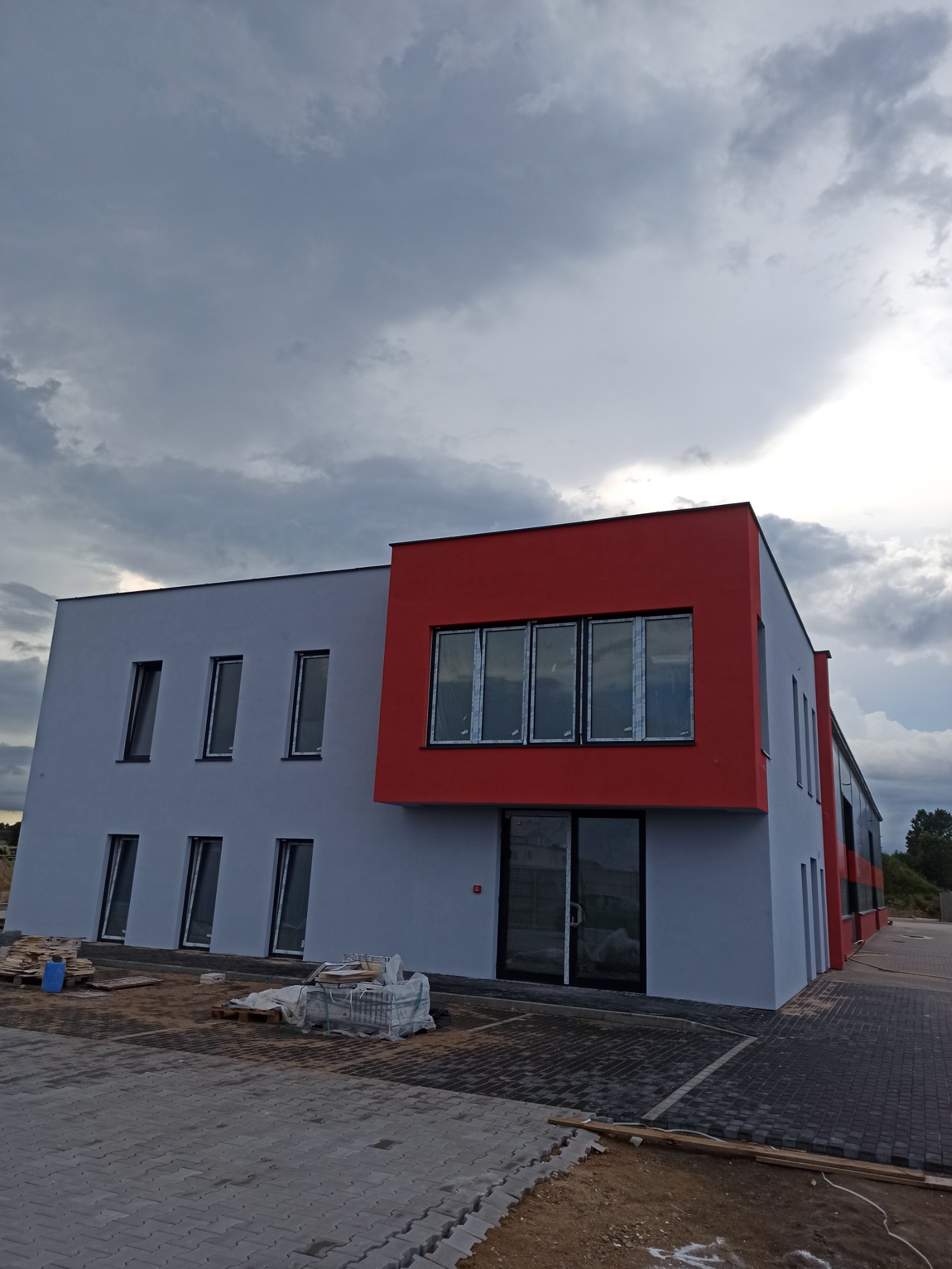 Nowo wybudowany budynek biurowy z szarą elewacją i czerwonym akcentem, z widocznymi oknami i terenem wokół w trakcie zagospodarowania.