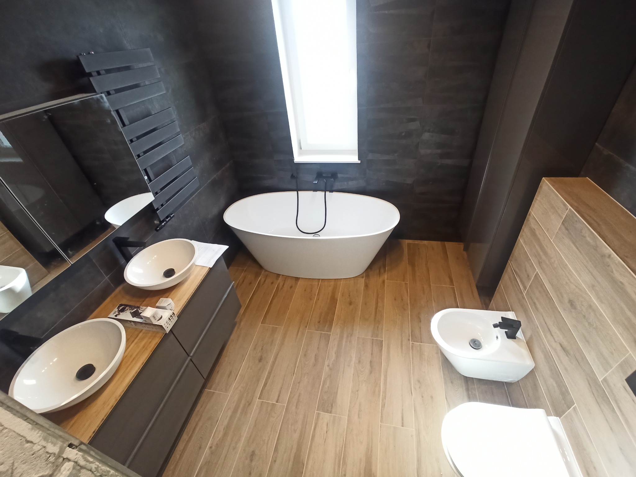 Nowoczesna łazienka z ciemnymi płytkami imitującymi kamień, wolnostojącą białą wanną pod oknem, drewnopodobną podłogą, umywalkami nablatowymi i czarną grzejnikową drabinką.