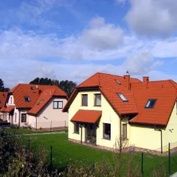 Rząd nowych, parterowych dom&oacute;w jednorodzinnych z czerwonymi dachami, widocznych za niskim ogrodzeniem z siatki, na tle błękitnego nieba z chmurami.