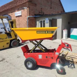 Czerwony, elektryczny wózek podnośnikowy z żółtą platformą załadunkową HG E1000, prezentowany w Witoszycach na tle budynku z cegły i wózka widłowego.