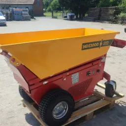 Elektryczny wózek transportowy HG E1000 z żółtą skrzynią ładunkową, stojący na palecie na placu budowy, w tle zaparkowane samochody i składowane materiały budowlane.