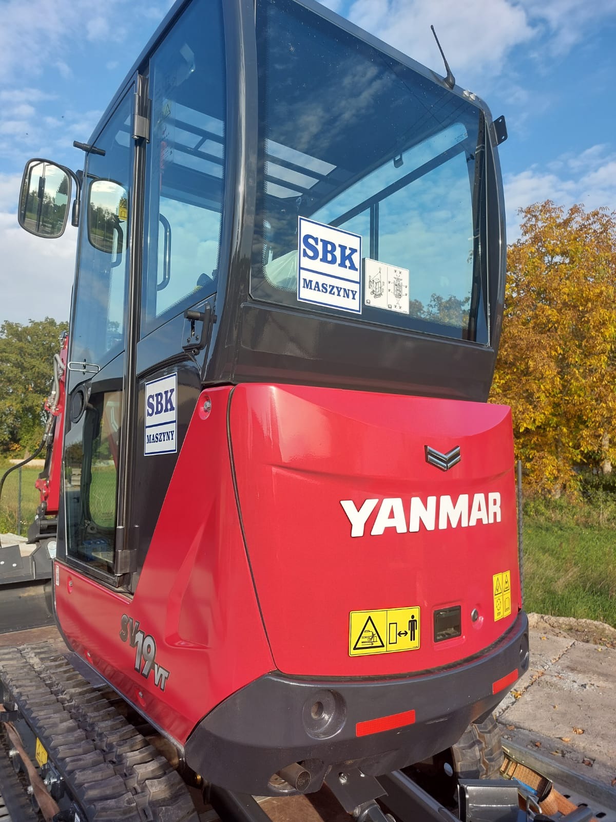 Czerwona, fabrycznie nowa maszyna Yanmar SV19VT z naklejkami SBK Maszyny na kabinie, stojąca na przyczepie w słoneczny dzień.
