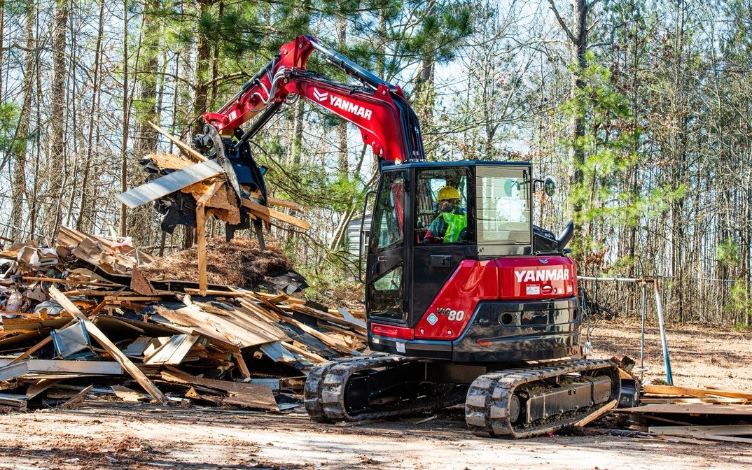 Czerwona maszyna Yanmar z operatorem w żółtym kasku i zielonej kamizelce, zgarnia gruz z budowy w lesie, widoczne drzewa w tle.
