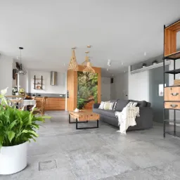 Przestronne, jasne wnętrze mieszkania z szarą sofą, drewnianym stołem kawowym i regałem z elementami drewna, oświetlone trzema ażurowymi lampami wiszącymi.