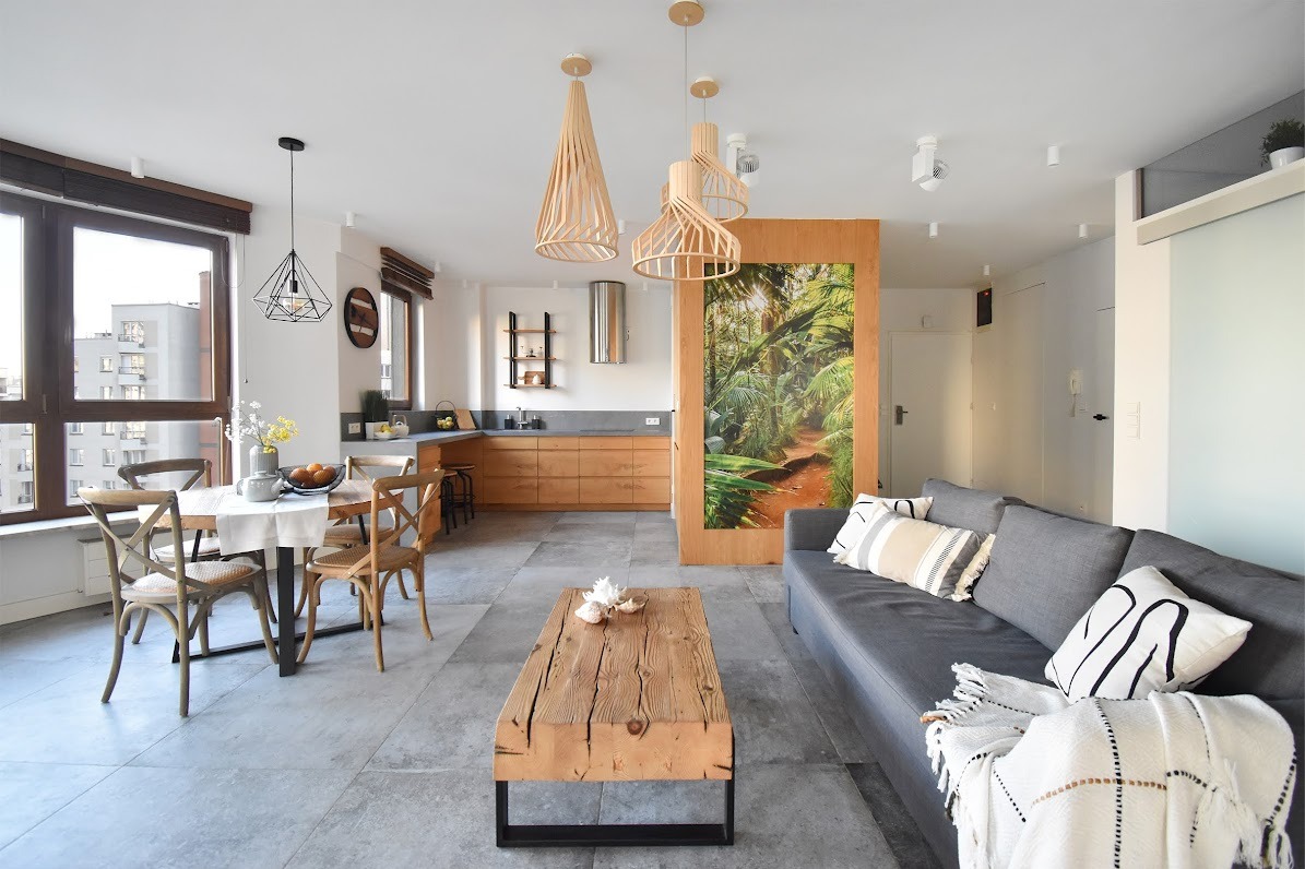 Jasne wnętrze mieszkania z drewnianą podłogą, stołem jadalnym z krzesłami, szarą sofą i drewnianym stolikiem kawowym, oświetlone wiszącymi lampami o ażurowych abażurach oraz z fototapetą z motywem...