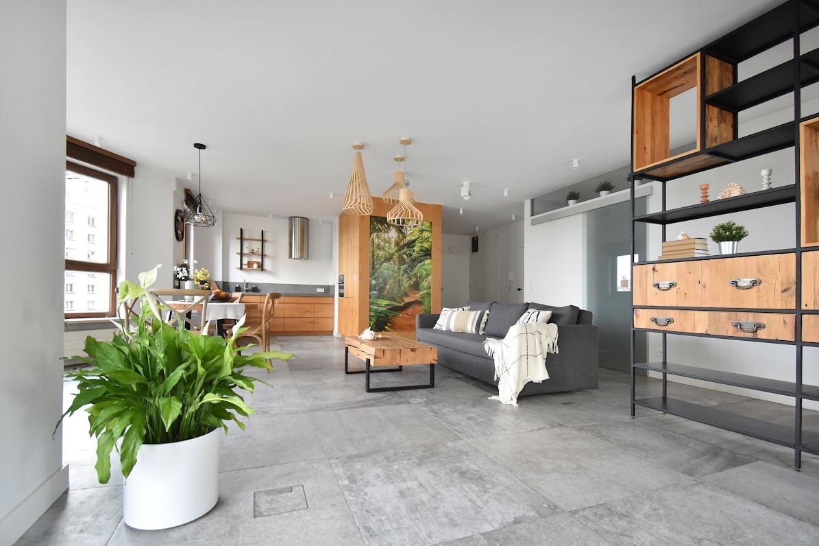 Przestronne, jasne wnętrze mieszkania z szarą sofą, drewnianym stołem kawowym i regałem z elementami drewna, oświetlone trzema ażurowymi lampami wiszącymi.