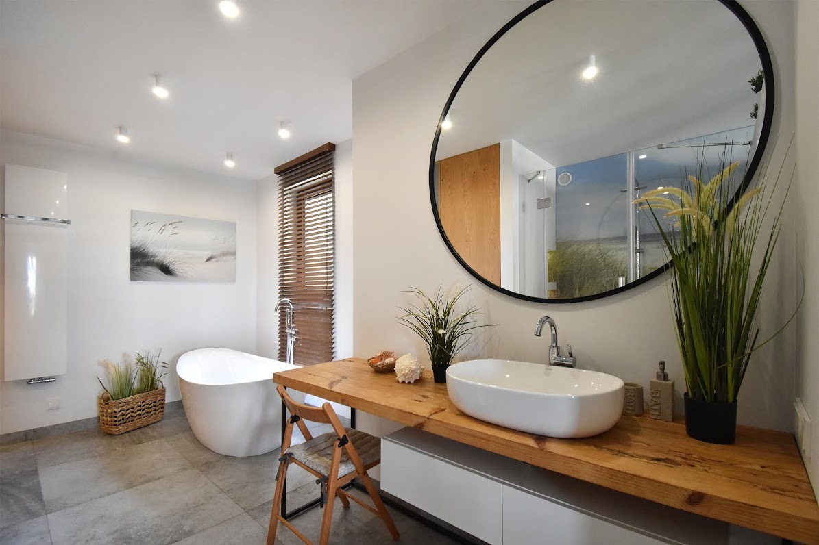 Nowoczesna łazienka z wolnostojącą wanną, drewnianym blatem pod umywalką i okrągłym lustrem, uzupełniona dekoracjami z motywem plaży.