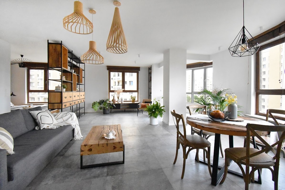 Jasne, przestronne wnętrze mieszkania z drewnianymi lampami wiszącymi, szarą sofą i drewnianym stolikiem kawowym na tle dużych okien.