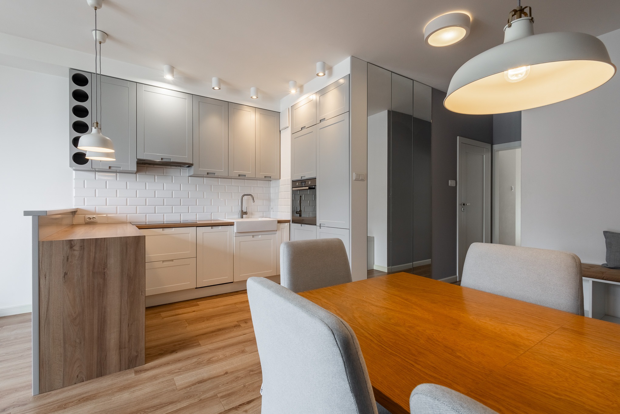 Jasna kuchnia z drewnianym stołem i szarymi krzesłami. Białe szafki, drewniany blat, lampy wiszące. Nowoczesne wnętrze z elementami retro.
