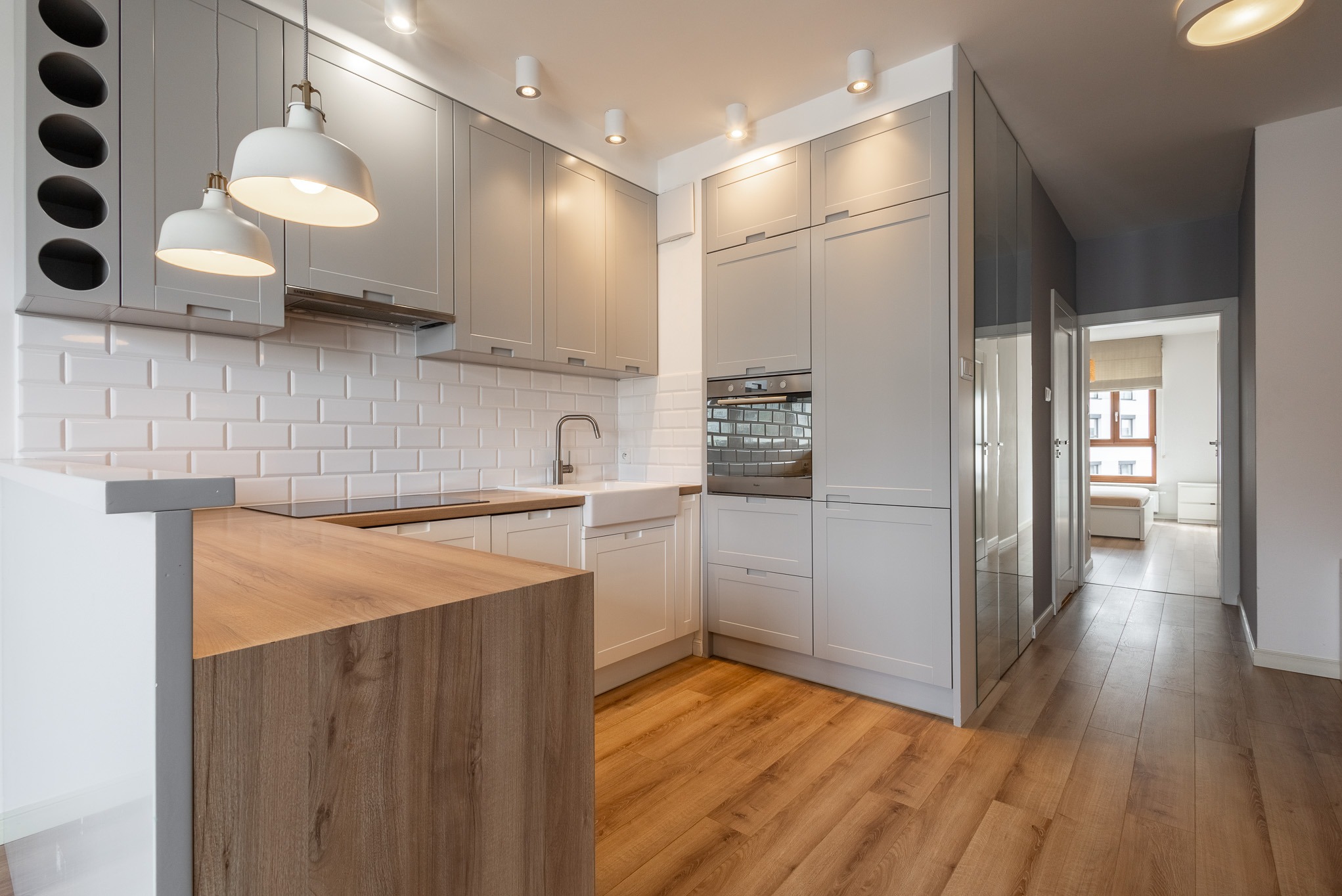 Jasna kuchnia w stylu skandynawskim: białe płytki, drewniany blat, szare szafki, lampy wiszące. Widok na pokój przez otwarte drzwi, parkiet na podłodze.