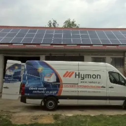 Furgonetka firmy Hymon z grafiką paneli słonecznych zaparkowana przed budynkiem gospodarczym z zamontowaną instalacją fotowoltaiczną na dachu.