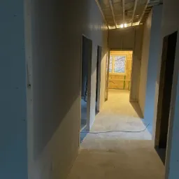 Korytarz w trakcie remontu z widoczną konstrukcją sufitu, ścianami przygotowanymi do dalszych prac wykończeniowych i oknem w oddali.