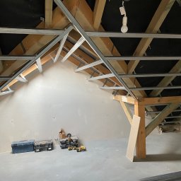 StanBud Michał Stanek - Poddasze w trakcie adaptacji: drewniana konstrukcja dachu z zamontowanymi metalowymi profilami, przygotowanymi pod montaż okładzin. Widoczne narzędzia i materiały budowlane. Ściany wstępnie...