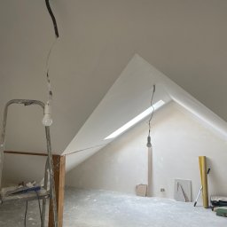 StanBud Michał Stanek - Poddasze w trakcie prac wykończeniowych: ściany pokryte świeżą warstwą, widoczne narzędzia na drabinie, lampa wisząca z sufitu, okno dachowe w skośnej ścianie. Podłoga z widocznymi śladami prac.