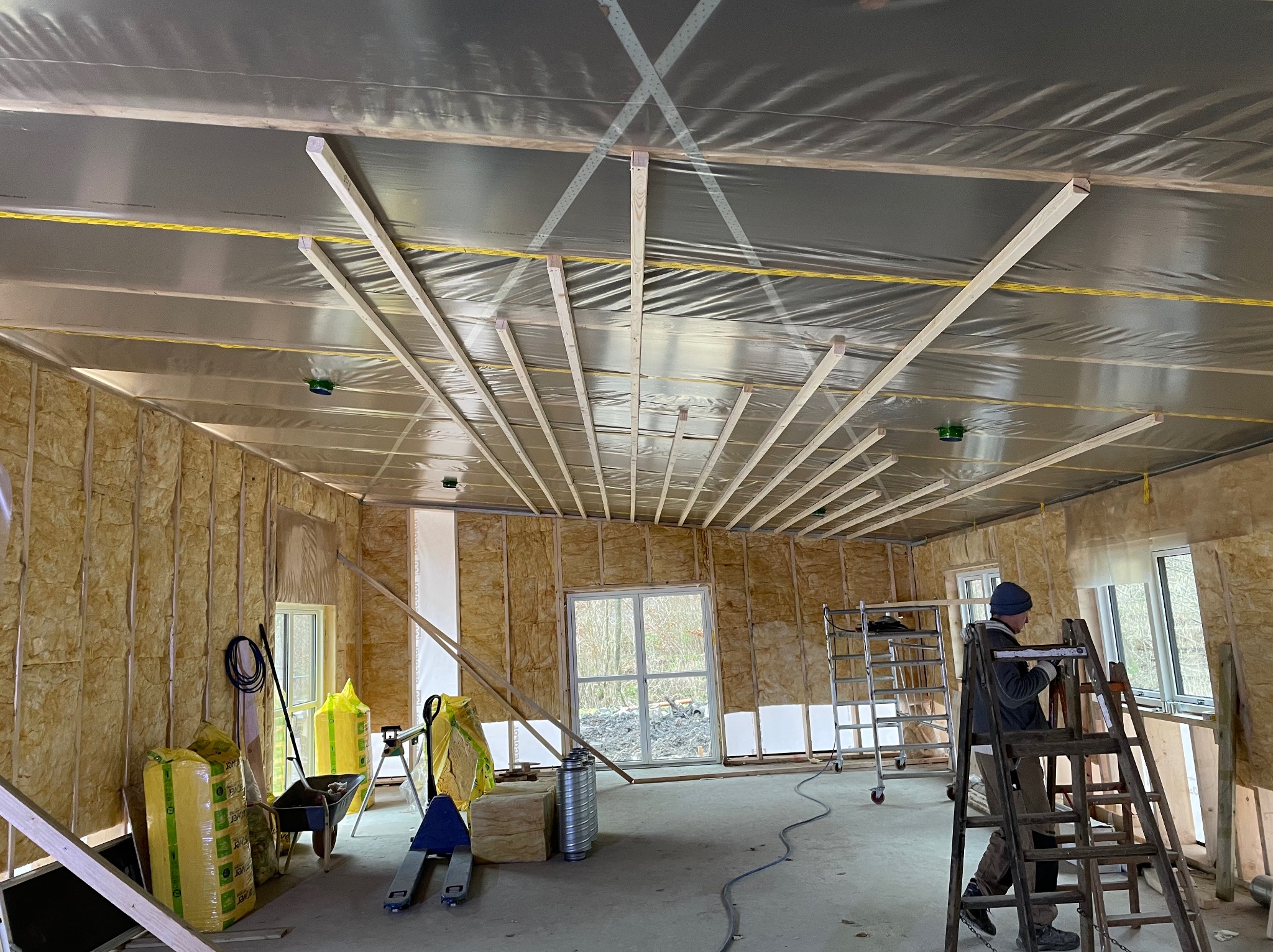 Wnętrze w trakcie prac wykończeniowych: izolacja ścian wełną mineralną, folia paroizolacyjna na suficie i drewniane profile pod konstrukcję sufitu podwieszanego, pracownik na drabinie.