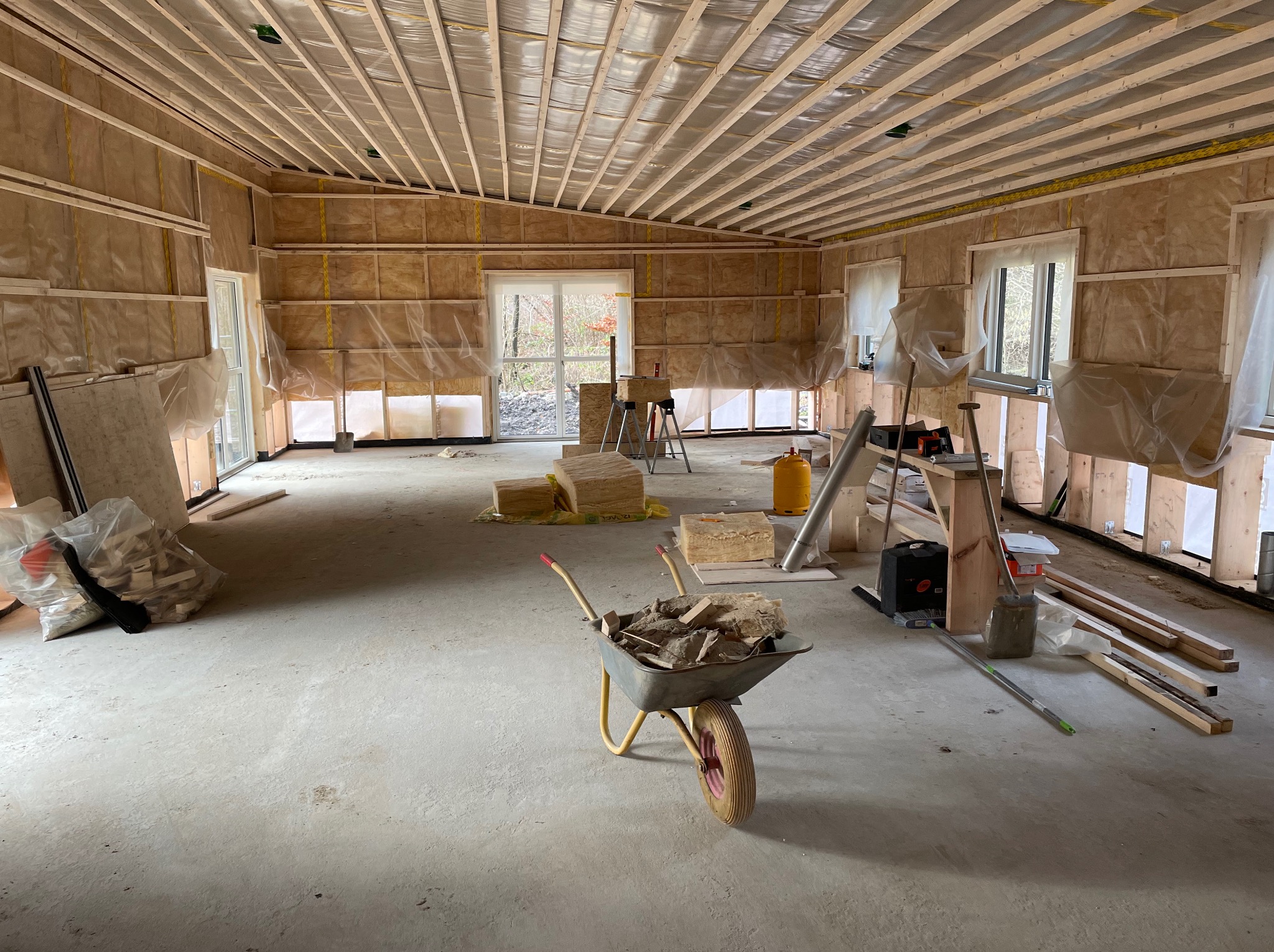 Wnętrze w trakcie prac wykończeniowych: widoczne ocieplone ściany z folią, drewniana konstrukcja sufitu, taczka z gruzem na betonowej podłodze, okna z widokiem na zewnątrz.