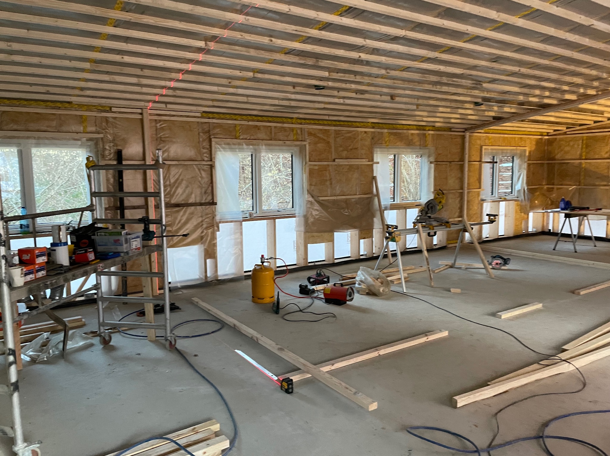 Wnętrze w trakcie remontu w Rzeszowie: widoczne ocieplone ściany z folią, drewniana konstrukcja sufitu, narzędzia, drabina i laserowy niwelator wyznaczający poziom.