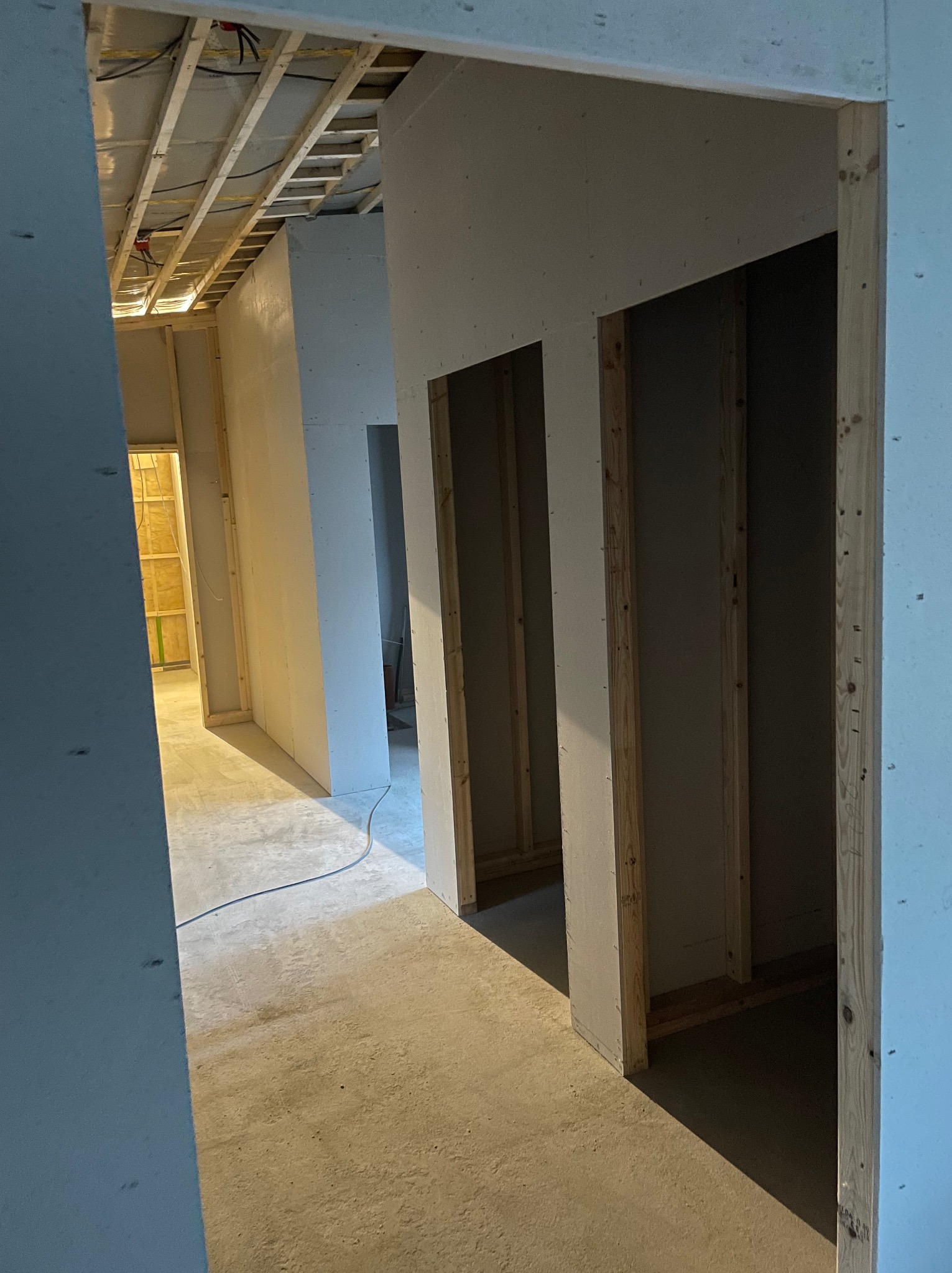 Wnętrze w trakcie budowy z widocznymi ścianami z płyt kartonowo-gipsowych, drewnianą konstrukcją sufitu z instalacją elektryczną oraz surową betonową podłogą, widziane z perspektywy przejścia.