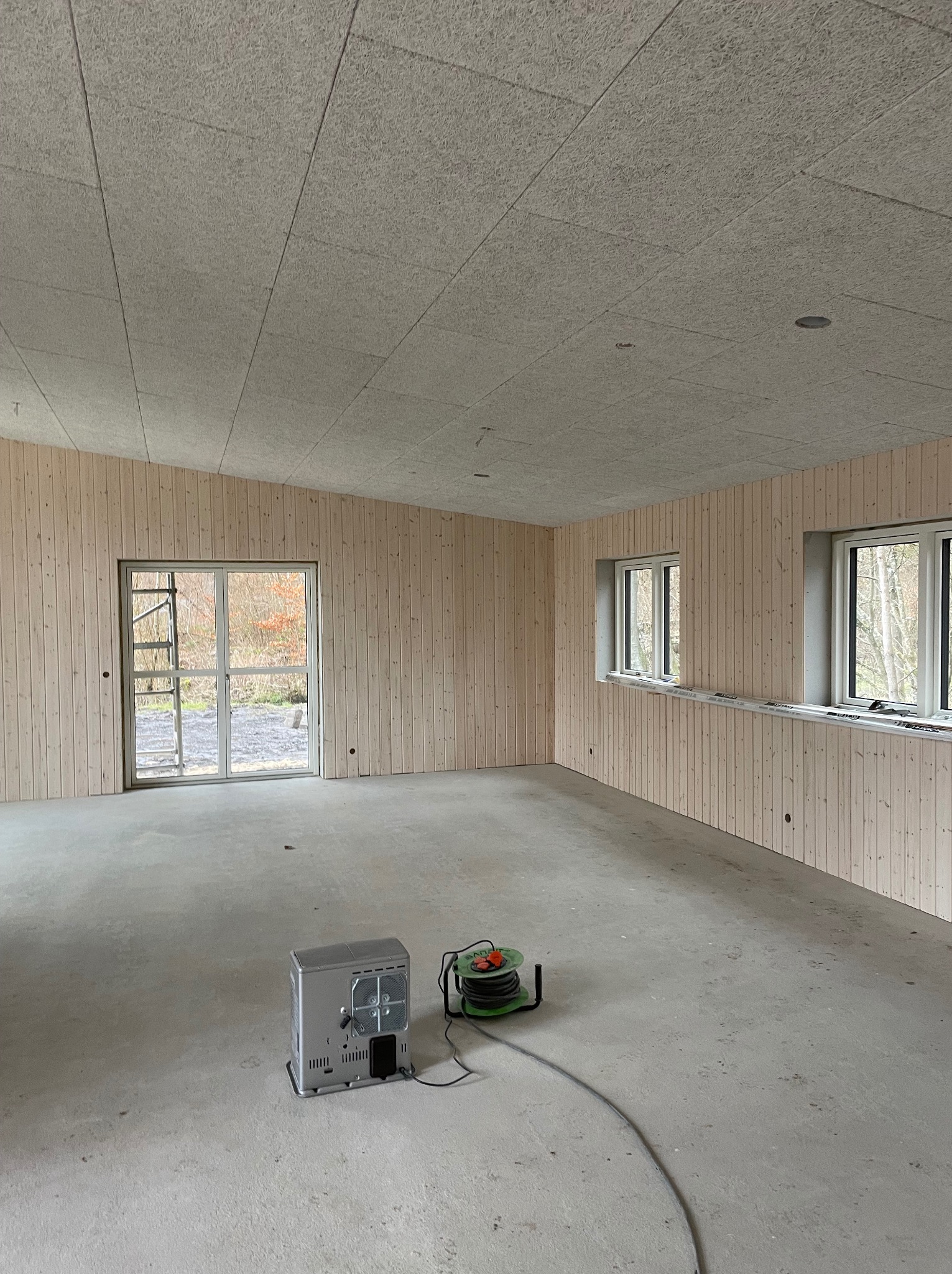 Surowe wnętrze w trakcie wykańczania: ściany z boazerii, sufit z płyt o wyraźnej fakturze, betonowa podłoga z grzejnikiem i zwiniętym przedłużaczem, widok na zewnątrz przez przeszklone drzwi.