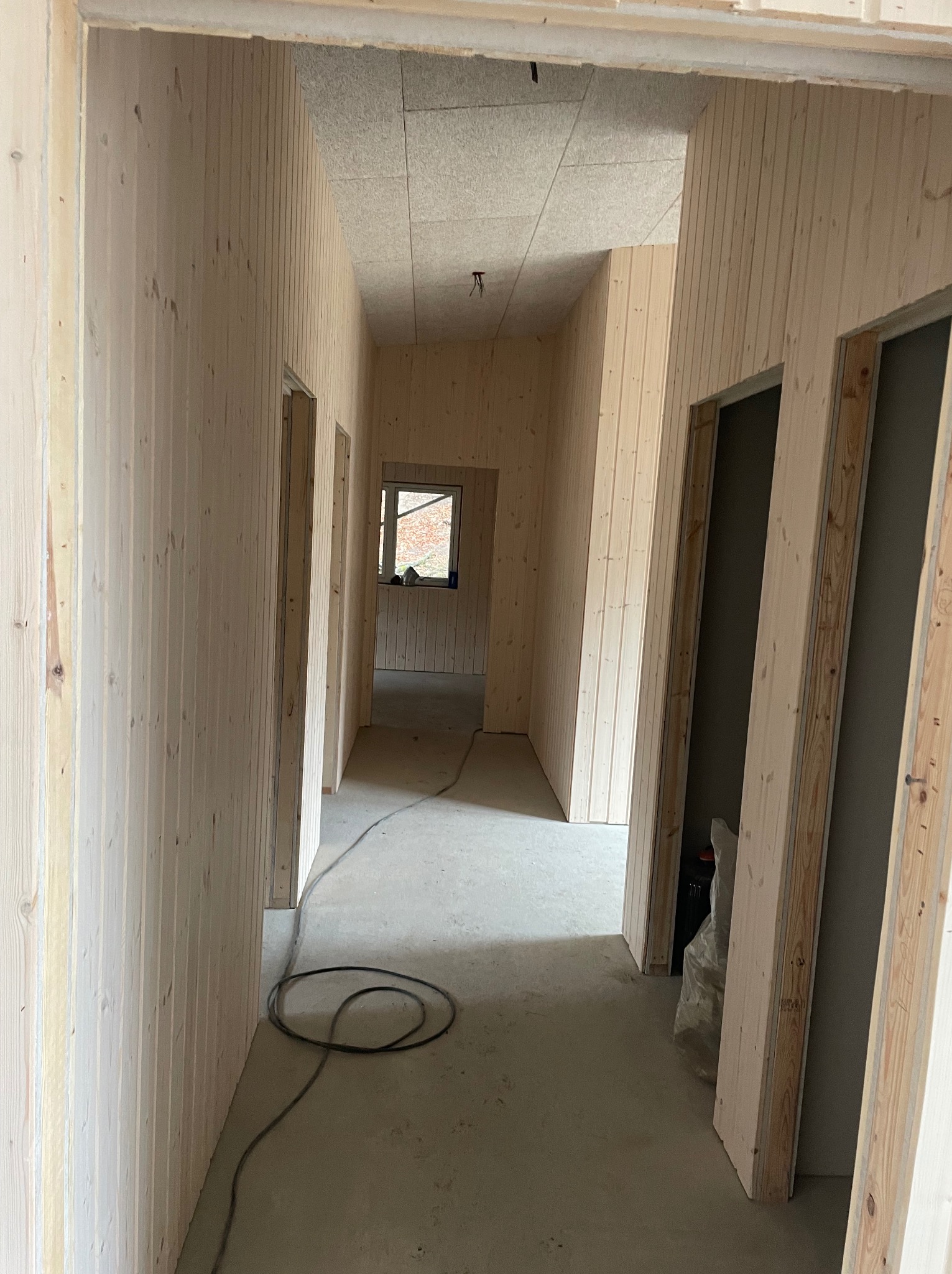 Korytarz w trakcie wykańczania, ściany i sufit wyłożone jasnymi panelami boazeryjnymi, widoczne kable na podłodze i surowe ościeżnice drzwiowe.