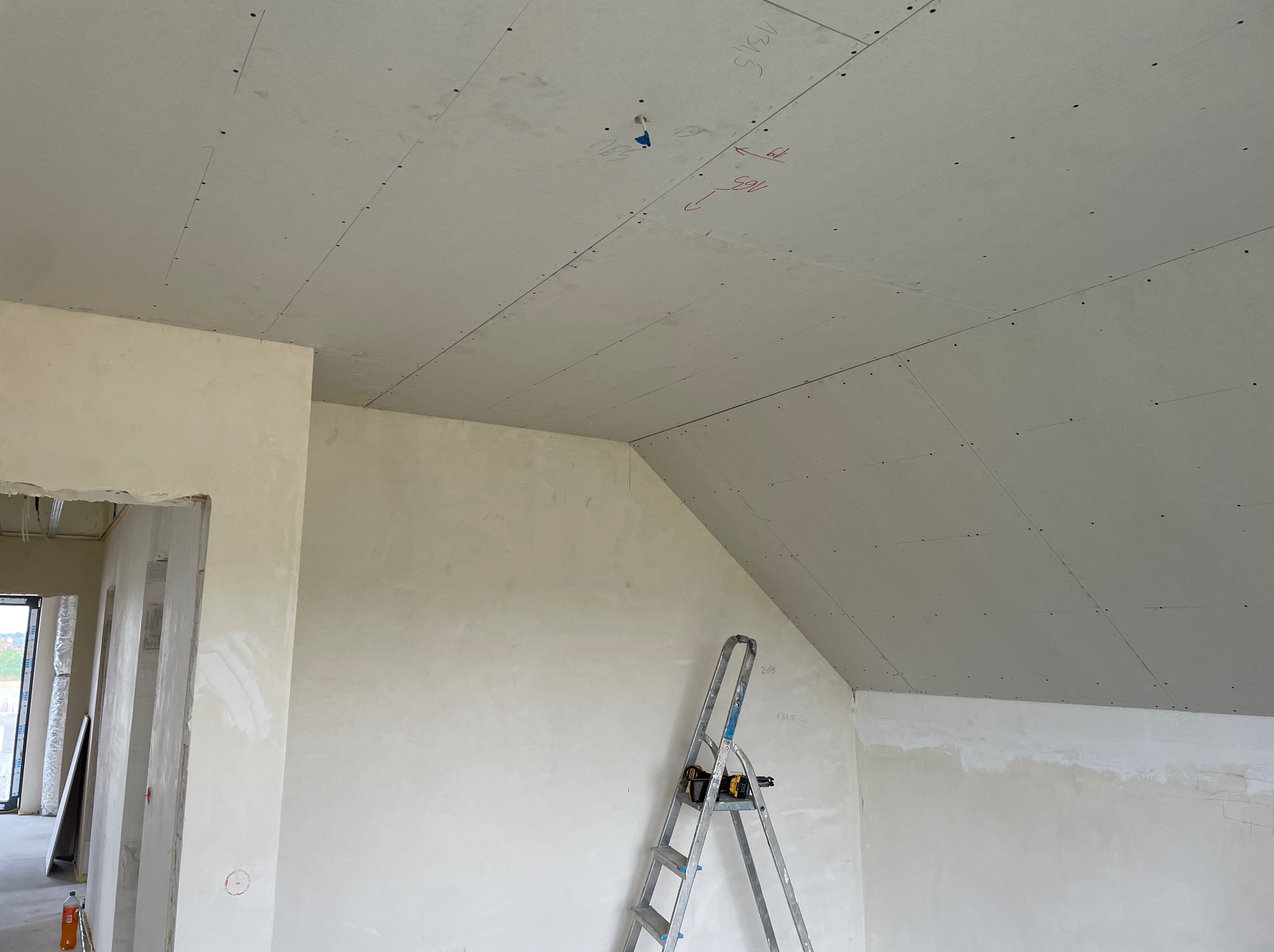 Wnętrze w trakcie wykończenia: sufit z płyt kartonowo-gipsowych z widocznymi śladami po wkrętach i notatkami, ściany wstępnie przygotowane pod malowanie, drabina aluminiowa oparta o ścianę.